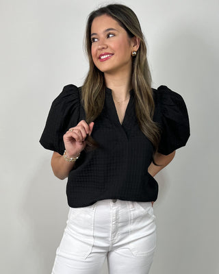 Model wearing a black puff sleeve blouse with a split neckline, styled with high-waisted white wide leg pants and clear slide sandals.