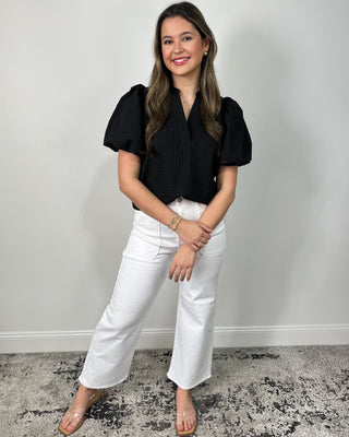 Model wearing a black puff sleeve blouse with a split neckline, styled with high-waisted white wide leg pants and clear slide sandals.