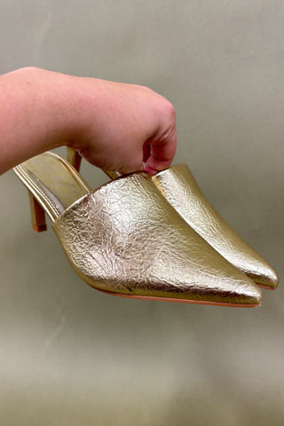 Gold high-heeled shoes held by a hand against a neutral background