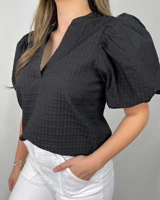 Model wearing a black puff sleeve blouse with a split neckline, styled with high-waisted white wide leg pants and clear slide sandals.