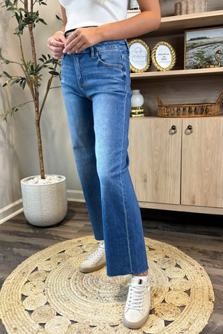 Person wearing blue jeans and white sneakers standing on a woven rug in a room with wooden furniture and decor.