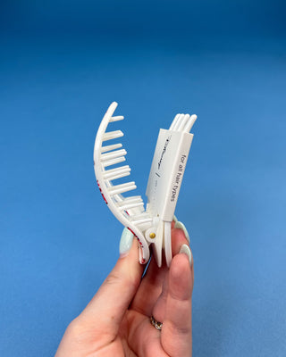 A hand holding a white plastic claw hair clip against a solid blue background. The clip is shown open, displaying its teeth and a white paper insert with black text that reads 'for all hair types'.