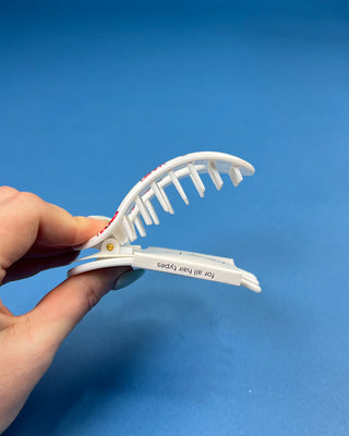 A hand holding a white plastic hair clipper against a solid blue background. The clipper has red and white printed text on its inner surface.