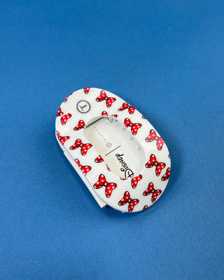 A white plastic hair clip featuring a red and white polka dot Minnie Mouse bow pattern, displayed on a solid blue background. The Disney brand name is visible on the front.