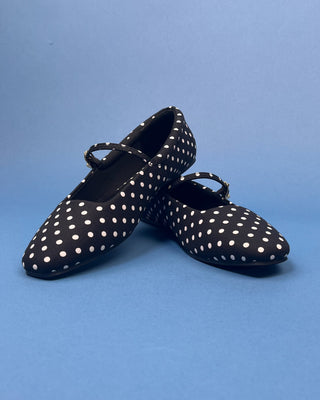 A pair of black Mary Jane flats with white polka dots, displayed on a solid blue background.