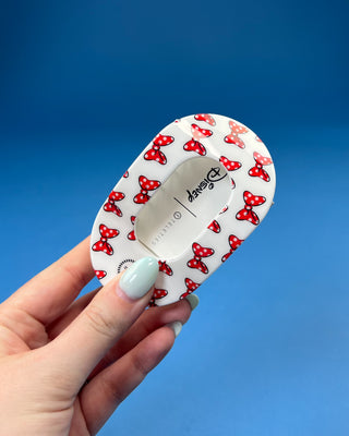 A hand with mint green nail polish holds a white oval plastic hair clip with a red and white polka dot bow pattern, featuring the Disney logo on the inside.