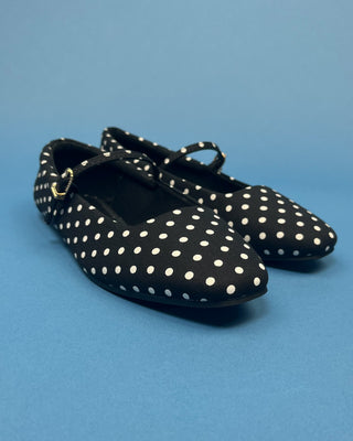 A pair of black ballet flats with white polka dots and a small gold-tone buckle detail on the side, displayed on a solid blue background.