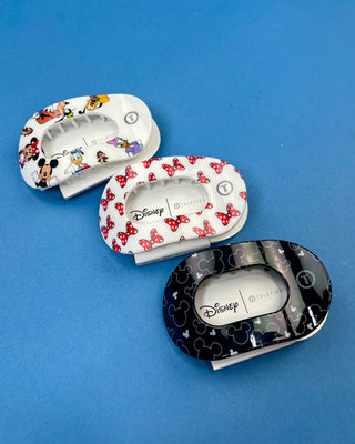 Three oval hair claw clips from Disney's Collection are arranged diagonally on a solid blue background. The top-left clip is white with colorful cartoon character prints, the middle clip is white with red bow patterns, and the bottom-right clip is black with silver Mickey Mouse head silhouettes.