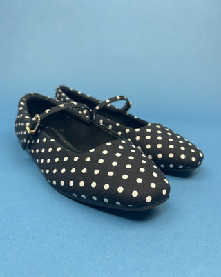 A pair of black ballet flats with white polka dots and a small gold buckle on the strap, set against a solid blue background.