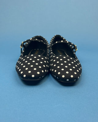 A pair of black Mary Jane style shoes with white polka dots and small gold buckle straps, photographed against a solid blue background.
