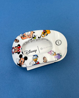 A hand holding a white Disney Teleties hair clip decorated with colorful cartoon characters like Mickey Mouse, Minnie Mouse, Donald Duck, and Daisy Duck, set against a solid blue background.
