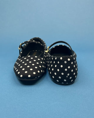 A pair of black Mary Jane style shoes with white polka dots, featuring a small gold buckle on the strap, set against a solid blue background.