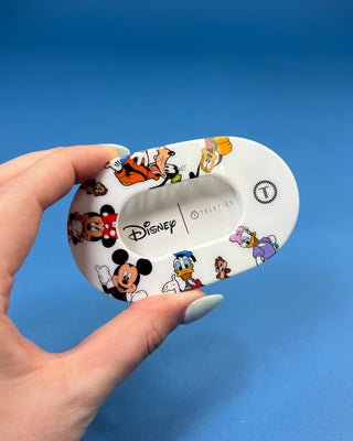 A hand with light blue nail polish holds a white oval hair clip decorated with colorful Disney character illustrations, including Mickey Mouse, Minnie Mouse, Donald Duck, Daisy Duck, Goofy, Pluto, and Tinker Bell, against a solid blue background.