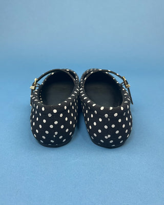 A pair of black Mary Jane style shoes with white polka dots and small gold buckle details, shown from the back on a solid blue background.
