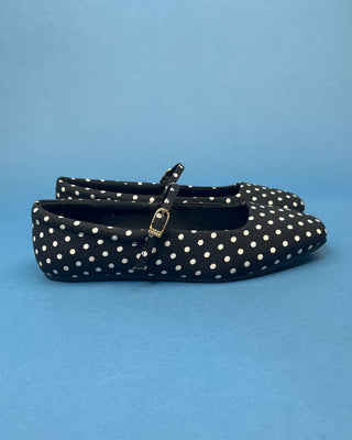 A pair of black flat shoes with white polka dots and a small gold-tone buckle detail, set against a solid blue background.