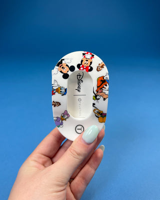 A hand holding a white Disney Teleties hair clip decorated with colorful cartoon characters like Mickey Mouse, Minnie Mouse, Donald Duck, and Daisy Duck, set against a solid blue background.