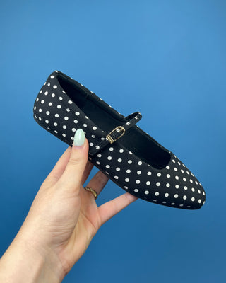 A hand with mint green nail polish holds a black and white polka dot Mary Jane flat shoe against a solid blue background.
