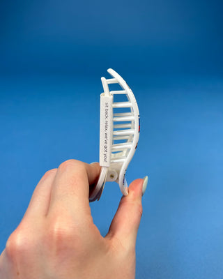 A hand holding a white plastic claw hair clip against a solid blue background. The clip features the printed text 'SIT DOWN, RELAX, WE'LL TEAR YOU.