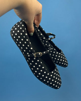 A pair of black Mary Jane shoes with white polka dots and a small gold buckle, held by a hand against a solid blue background.
