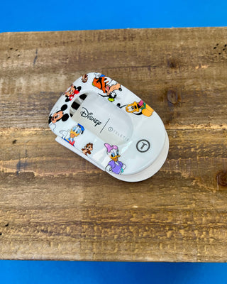 A hand holding a white Disney Teleties hair clip decorated with colorful cartoon characters like Mickey Mouse, Minnie Mouse, Donald Duck, and Daisy Duck, set against a solid blue background.
