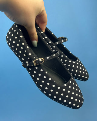 A hand with mint green nail polish holds a black and white polka dot Mary Jane flat shoe against a solid blue background.