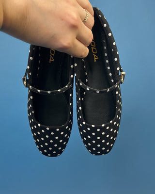A pair of black Mary Jane flats with white polka dots, held by a hand against a solid blue background. 