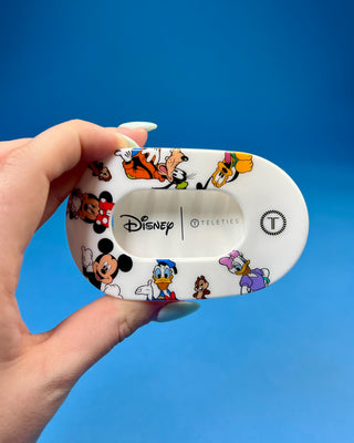 A hand holding a white Disney Teleties hair clip decorated with colorful cartoon characters like Mickey Mouse, Minnie Mouse, Donald Duck, and Daisy Duck, set against a solid blue background.