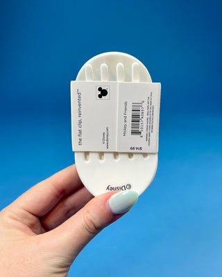 A hand holding a white plastic hair clipper against a solid blue background. The clipper has red and white printed text on its inner surface.