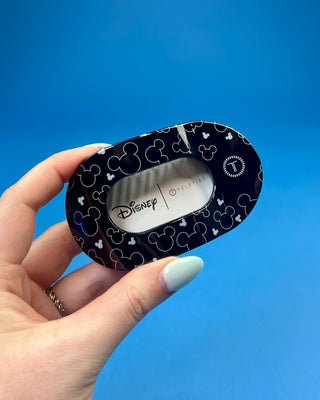 A hand holds a black hair claw clip against a solid blue background. The clip has a white paper label attached to it that reads 'for all hair types' in black text.