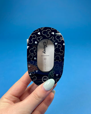 A hand holds a black hair claw clip against a solid blue background. The clip has a white paper label attached to it that reads 'for all hair types' in black text.