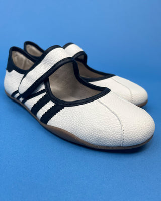 A pair of white pebbled leather Mary Jane shoes with black trim and three black stripes on the side, displayed on a solid blue background.