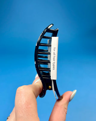 A hand holds a black hair claw clip against a solid blue background. The clip has a white paper label attached to it that reads 'for all hair types' in black text.