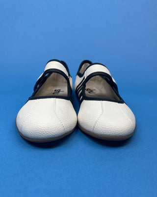 A pair of white leather Mary Jane shoes with black trim and straps, shown from the front on a solid blue background.