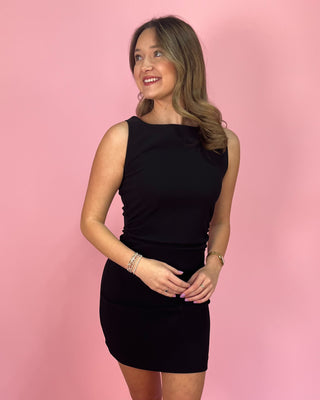 Black sleeveless bodycon mini dress with a high bateau neckline and fitted silhouette, styled with clear strap heels against a pink background.
