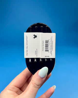 A hand holds a black hair claw clip against a solid blue background. The clip has a white paper label attached to it that reads 'for all hair types' in black text.