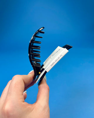 A hand holding a black plastic claw-style hair clip with a white paper tag attached, against a solid blue background.