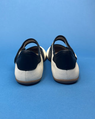A pair of women's Mary Jane shoes shown from the back, featuring a black leather backstrap and heel counter, a textured off-white pebbled leather midsole, and a brown gum sole, set against a solid blue background.