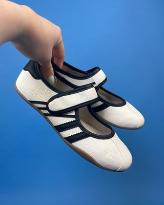 A pair of white pebbled leather Mary Jane shoes with black trim and three stripes, displayed on a solid blue background.