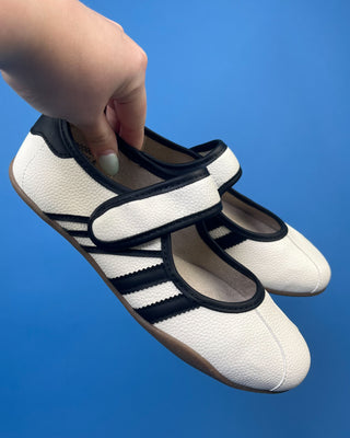 A hand holding a pair of white and black retro-style Mary Jane sneakers against a solid blue background.