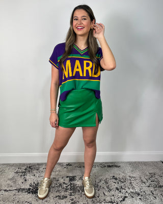 Model wearing a green faux leather mini skirt with a front slit, styled with a colorblock sweater vest and metallic gold sneakers.
