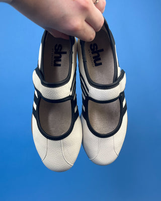A pair of white pebbled leather Mary Jane shoes with black trim and three stripes, displayed on a solid blue background.