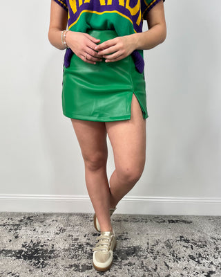 Model wearing a green faux leather mini skirt with a front slit, styled with a colorblock sweater vest and metallic gold sneakers.