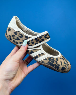 A hand holds a flat shoe with a leopard print pattern and cream-colored trim against a solid blue background.