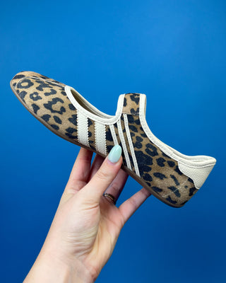 A leopard print slip-on shoe held by a hand against a solid blue background. The shoe features a tan and black leopard pattern with three white stripes on the side and a beige heel tab.