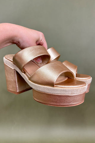 Gold platform sandals being held against a green background