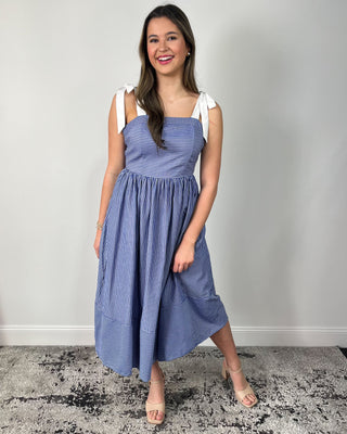 Model wearing a blue and white striped midi dress with bow tie shoulder straps, square neckline, gathered skirt, and side pockets, styled with nude heeled sandals.
