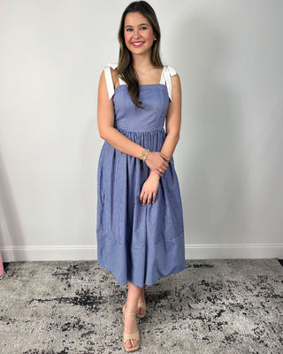 Model wearing a blue and white striped midi dress with bow tie shoulder straps, square neckline, gathered skirt, and side pockets, styled with nude heeled sandals.