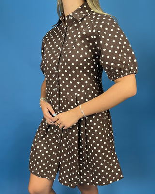 A woman wearing a brown short-sleeve button-front dress with white polka dots, standing against a solid blue background.
