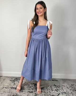 Model wearing a blue and white striped midi dress with bow tie shoulder straps, square neckline, gathered skirt, and side pockets, styled with nude heeled sandals.