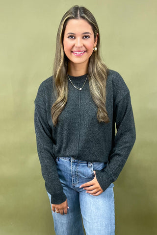 Woman wearing a dark gray sweater and light blue jeans against a green background